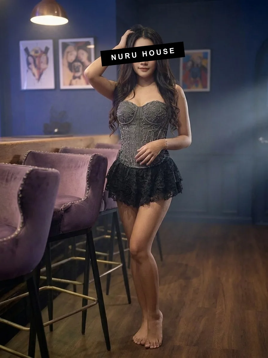 Standing barefoot by the velvet bar stools in a beaded grey corset and black lace skirt, one hand in her hair